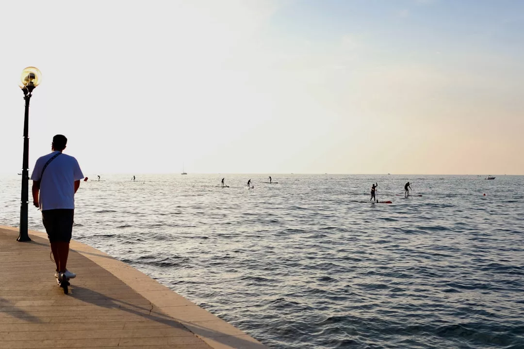 Una persona camina por un paseo marítimo de Mallorca mientras varias personas practican paddleboarding en un mar tranquilo y de lujo bajo un cielo luminoso.