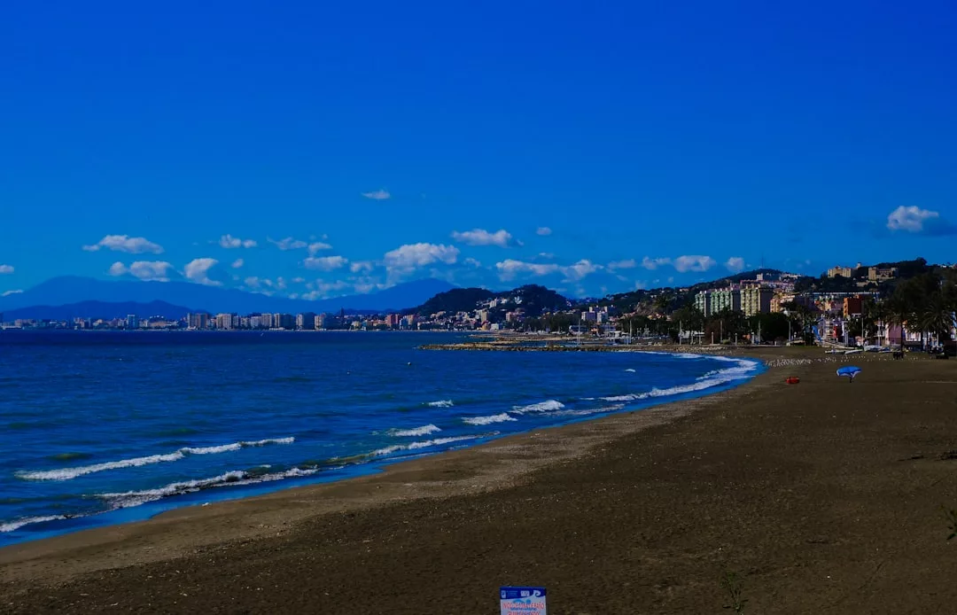 Una playa de arena se curva a lo largo de la costa con suaves olas, respaldada por una ciudad y montañas bajo un cielo azul claro, capturando el lujo de la impresionante costa de Mallorca.