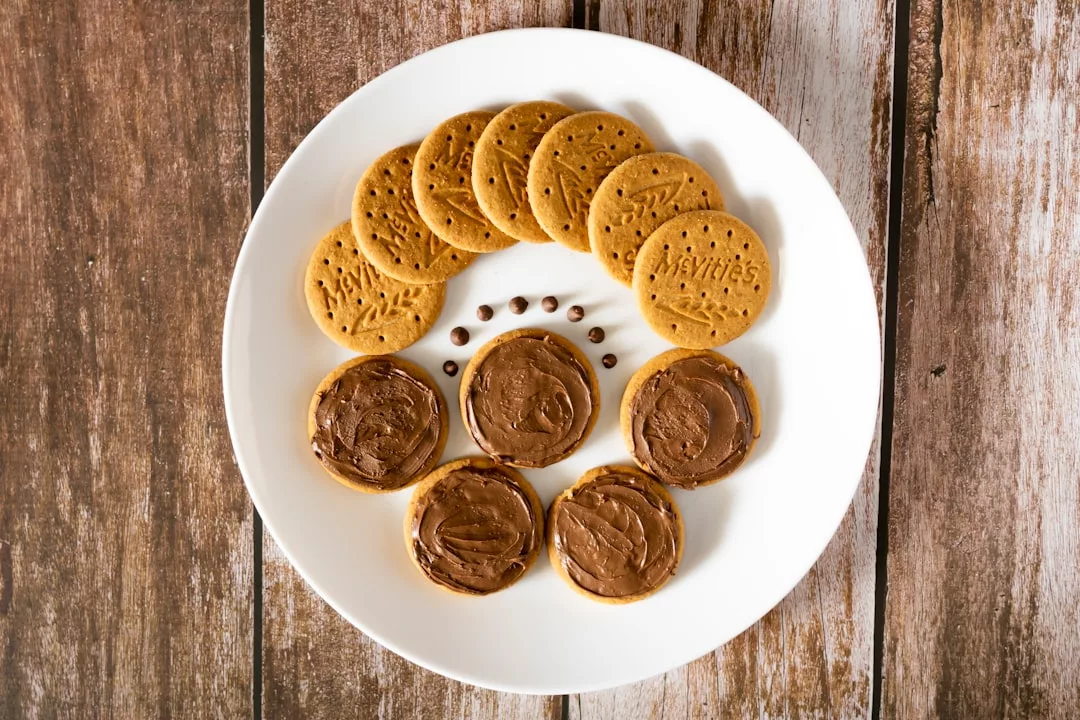 Un plato blanco con galletas redondas lisas y galletas cubiertas de chocolate para untar, dispuestas sobre una superficie de madera, aporta un toque de Lujo a tus mañanas mallorquinas.
