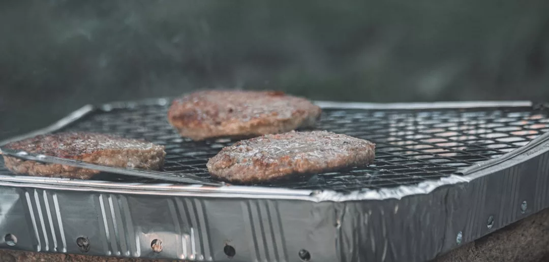 Tres hamburguesas se asan en una parrilla de aluminio desechable al aire libre, capturando el espíritu de un picnic informal de Lujo en la soleada Mallorca.