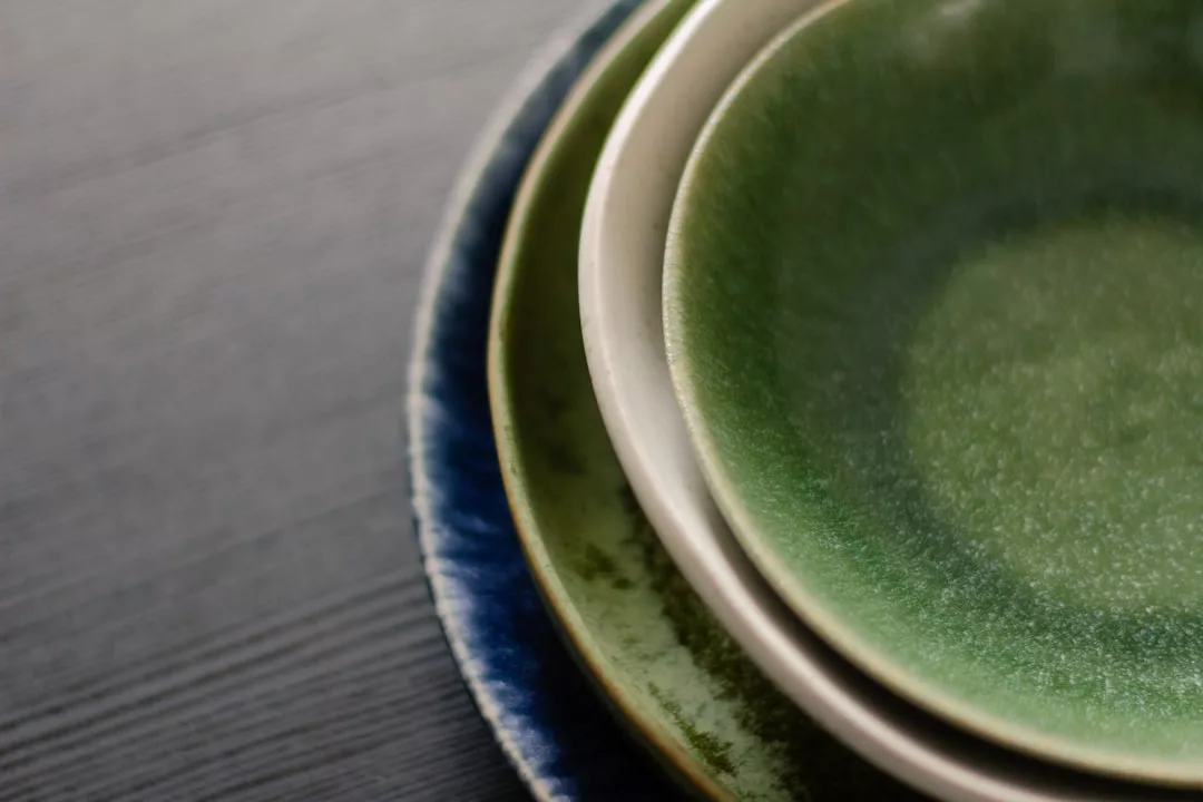 Primer plano de tres platos de cerámica apilados en tonos verde, crema y azul sobre una superficie de textura oscura, que evocan el relajado lujo de Mallorca.