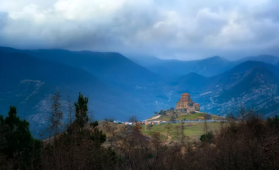 Una histórica iglesia de piedra se alza en lo alto de una colina de Mallorca, rodeada de montañas bajo un cielo nublado, con una hilera de coches aparcados cerca, añadiendo un toque de lujo al tranquilo paisaje.