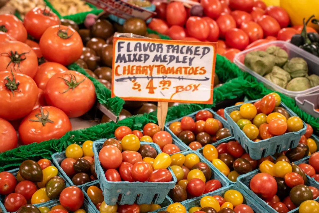 Un expositor de tomates cherry mezclados Lujo en cajas azules en un mercado, con un cartel que dice "Flavor Packed Mixed Medley Cherry Tomatoes $4.00 Box." Perfectos para llevar a su mesa los vibrantes sabores de Mallorca.