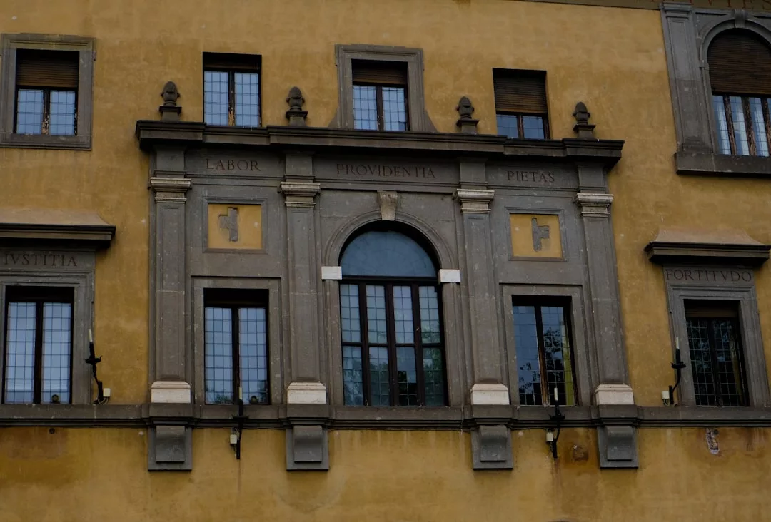 Sección de la fachada de un edificio amarillo de Mallorca con ventanas arqueadas y rectangulares, columnas y palabras en latín inscritas sobre las ventanas, que reflejan la singular mezcla de tradición y lujo de la región.