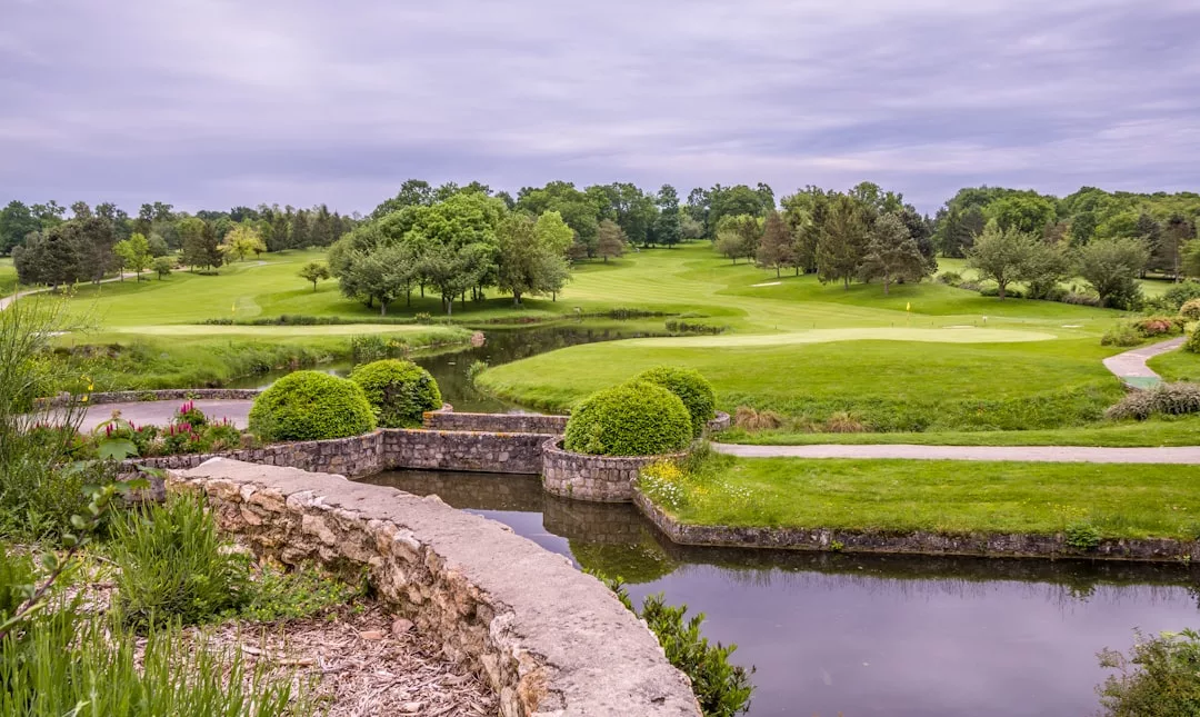 Un campo de golf ajardinado en Mallorca cuenta con césped cuidado, árboles, un pequeño estanque y un puente de piedra bajo un cielo nublado, ofreciendo un toque de lujo a cada ronda.