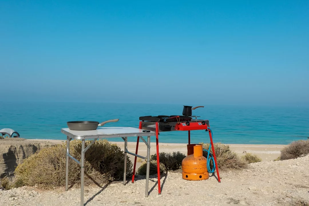 Una mesa plegable con una sartén y un hornillo de gas portátil con una olla están instalados en un acantilado de arena de Mallorca, con vistas al mar bajo un cielo azul despejado, creando un toque de lujo en medio de la naturaleza.