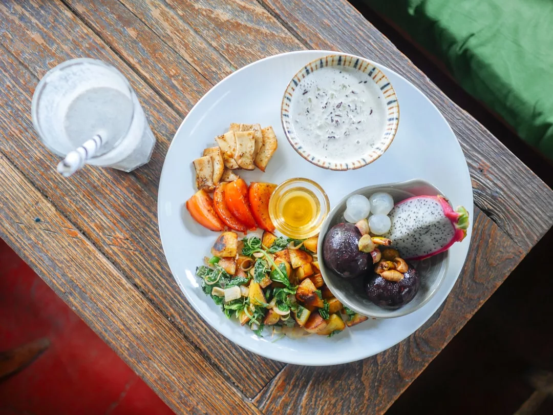 Un plato con verduras variadas, rodajas de tomate, tofu, un pequeño cuenco de yogur, fruta, frutos secos, miel y un vaso de agua con pajita sobre una mesa de madera ofrece una muestra del sencillo lujo de Mallorca en cada bocado.