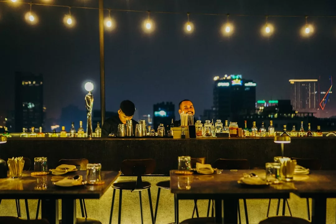 Dos camareros trabajan de noche en un bar al aire libre en Mallorca, con los edificios de la ciudad al fondo y las luces en lo alto. Las mesas en primer plano están preparadas para cenar, creando una escena de relajado lujo bajo las estrellas.
