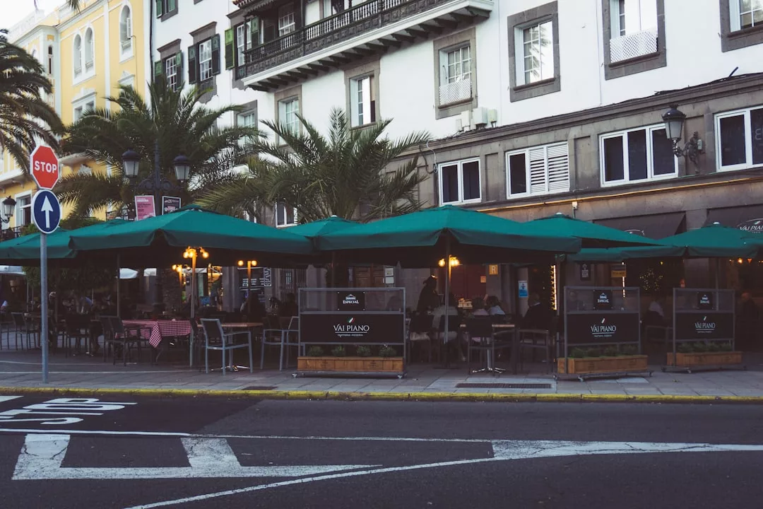 Vista de la calle de un restaurante al aire libre con sombrillas verdes, mesas y sillas en una acera de Mallorca, junto a un edificio con balcones y palmeras para darle un toque de lujo.