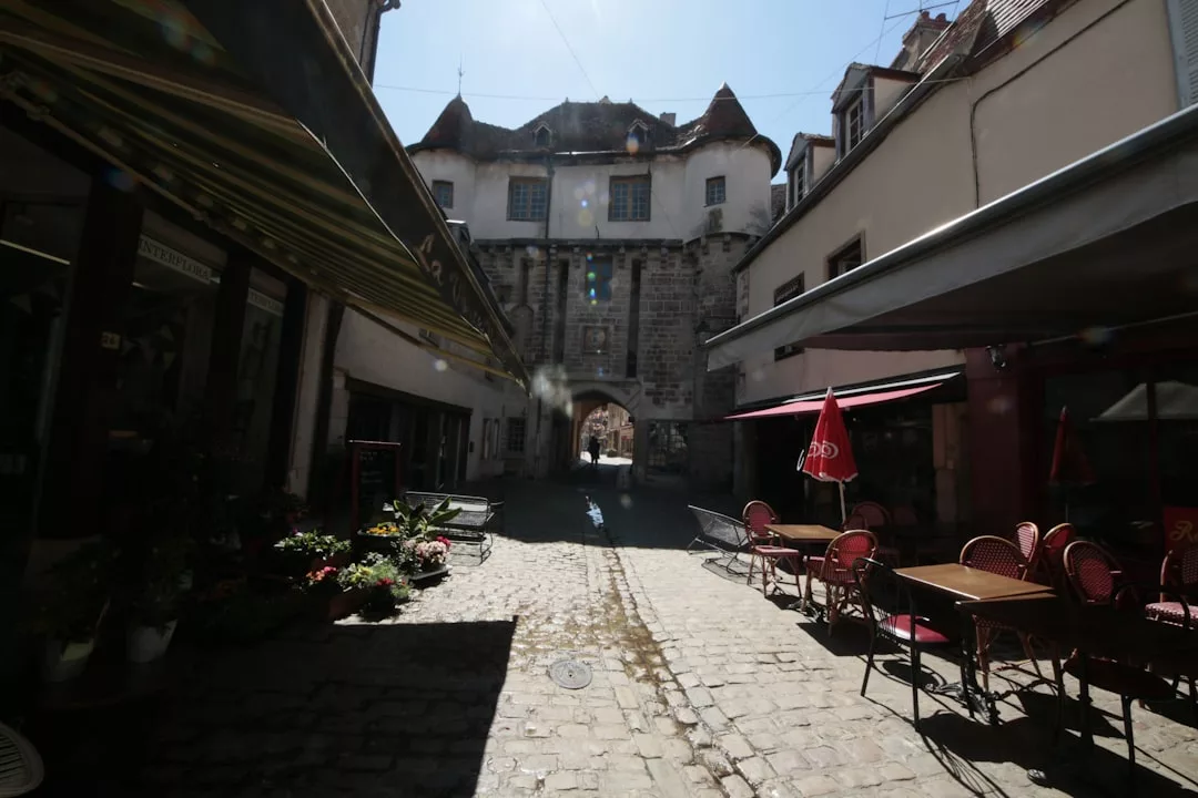 Una calle adoquinada con cafeterías al aire libre y tiendas conduce a un arco de piedra bajo un edificio histórico a plena luz del día, capturando el relajado lujo de Mallorca.