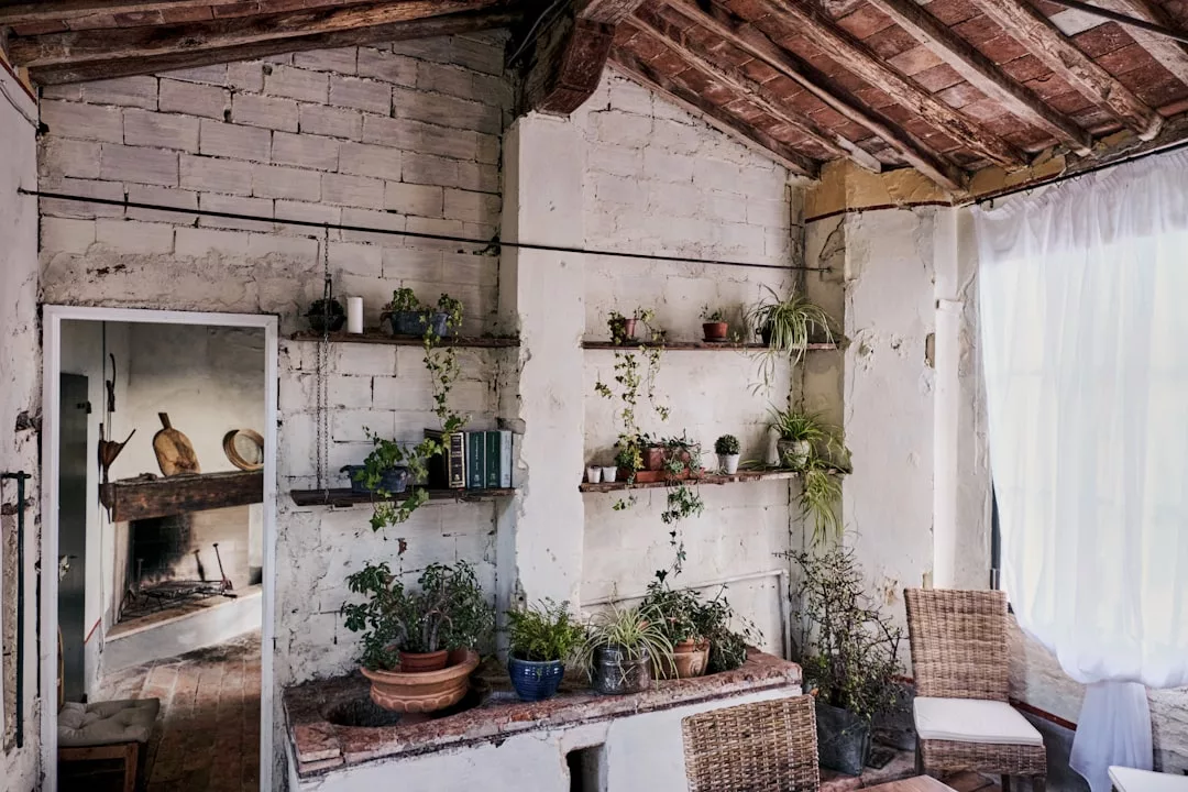 Habitación rústica con paredes de ladrillo encalado, estanterías de madera con plantas y libros, muebles de mimbre y la luz del sol filtrándose a través de una cortina, que evoca el lujo desenfadado de un escondite en Mallorca.
