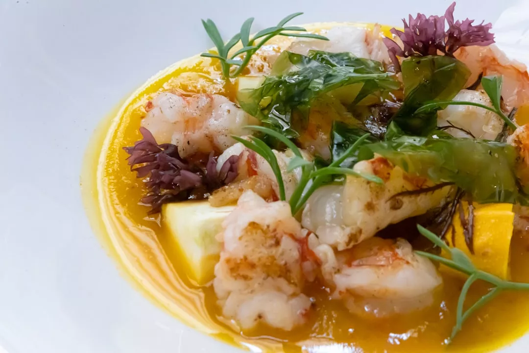 Un plato de marisco inspirado en Mallorca, con gambas, salsa amarilla, algas, hierbas frescas y guarniciones en un plato blanco para darle un toque de lujo.
