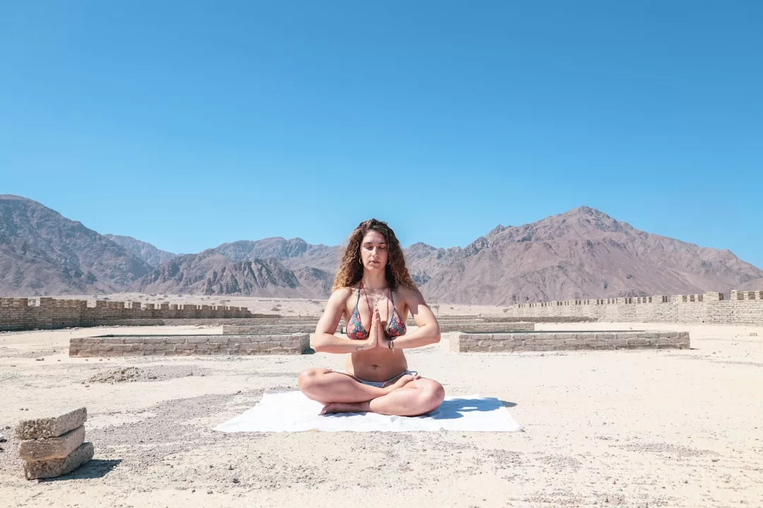Una mujer se sienta con las piernas cruzadas sobre una toalla blanca al aire libre, meditando con las manos en postura de oración frente a las impresionantes montañas y el cielo azul claro de Mallorca, disfrutando de un momento de lujo y serenidad.
