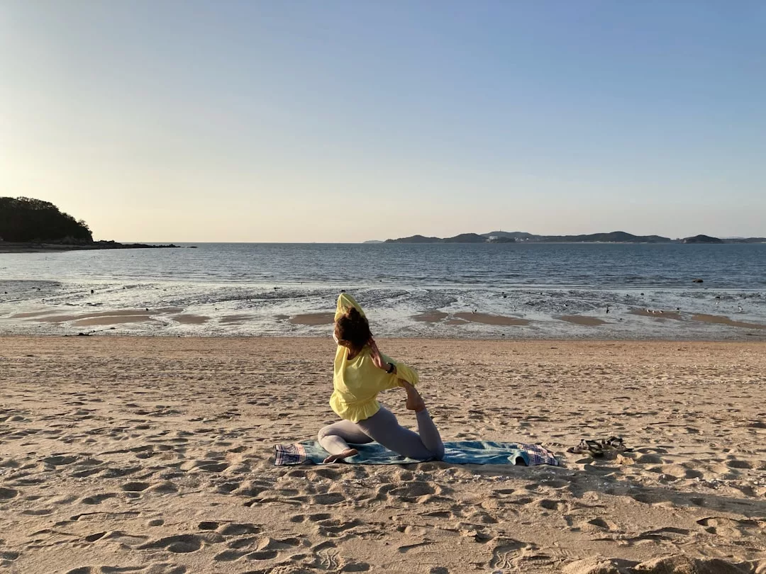 Una persona practica una postura de yoga sobre una esterilla en una playa de arena de Mallorca cerca del agua, con islas lejanas, un cielo despejado y un aire de lujo en el sereno fondo.