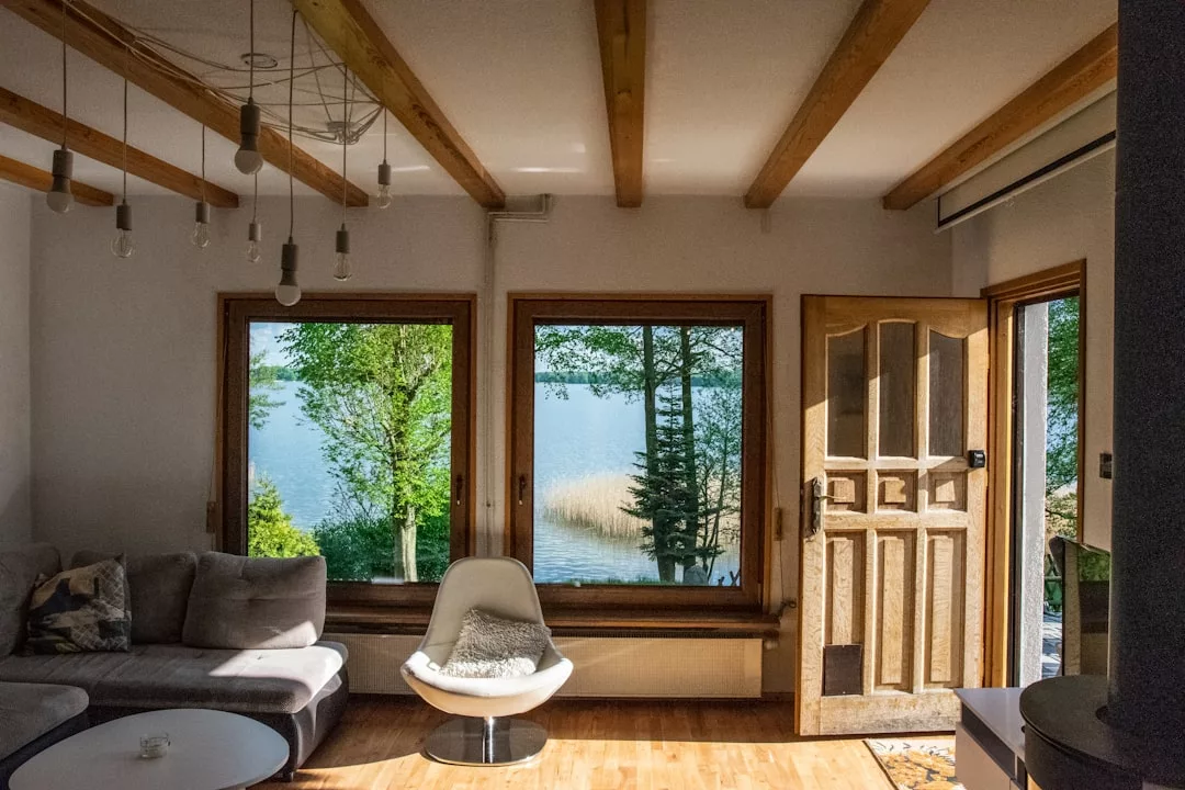 Un salón soleado en Mallorca con vigas de madera, un sofá, una silla blanca y grandes ventanales que ofrecen vistas de lujo de árboles y un lago; la puerta principal está abierta.