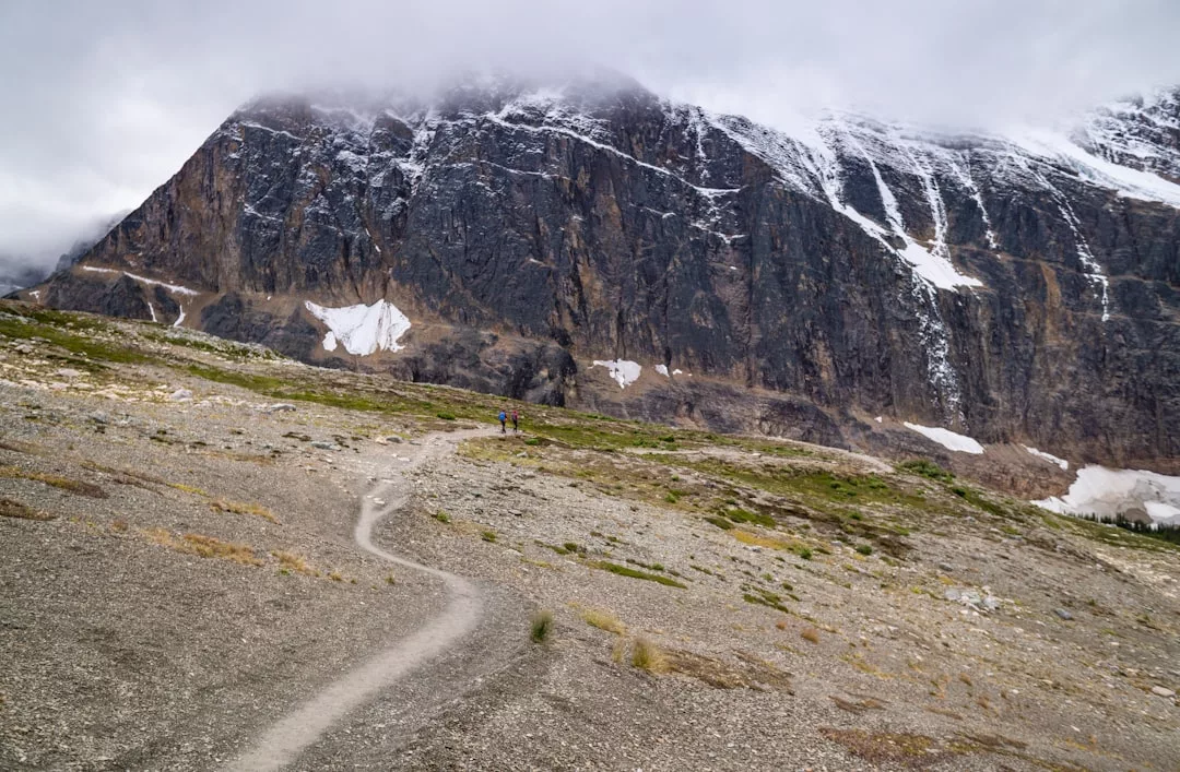Un sinuoso sendero de tierra conduce a una montaña rocosa, cubierta de nieve, que recuerda la belleza agreste de Mallorca, parcialmente velada por nubes bajas bajo un cielo encapotado: una joya oculta del lujo natural.