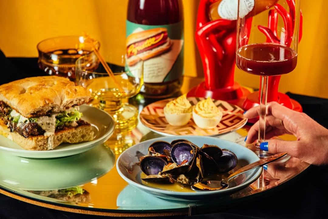 Un plato con un bocadillo, un cuenco de mejillones morados, huevos endiablados y bebidas se disponen sobre una superficie espejada en Lujo Mallorca, con una mano sosteniendo un vaso.