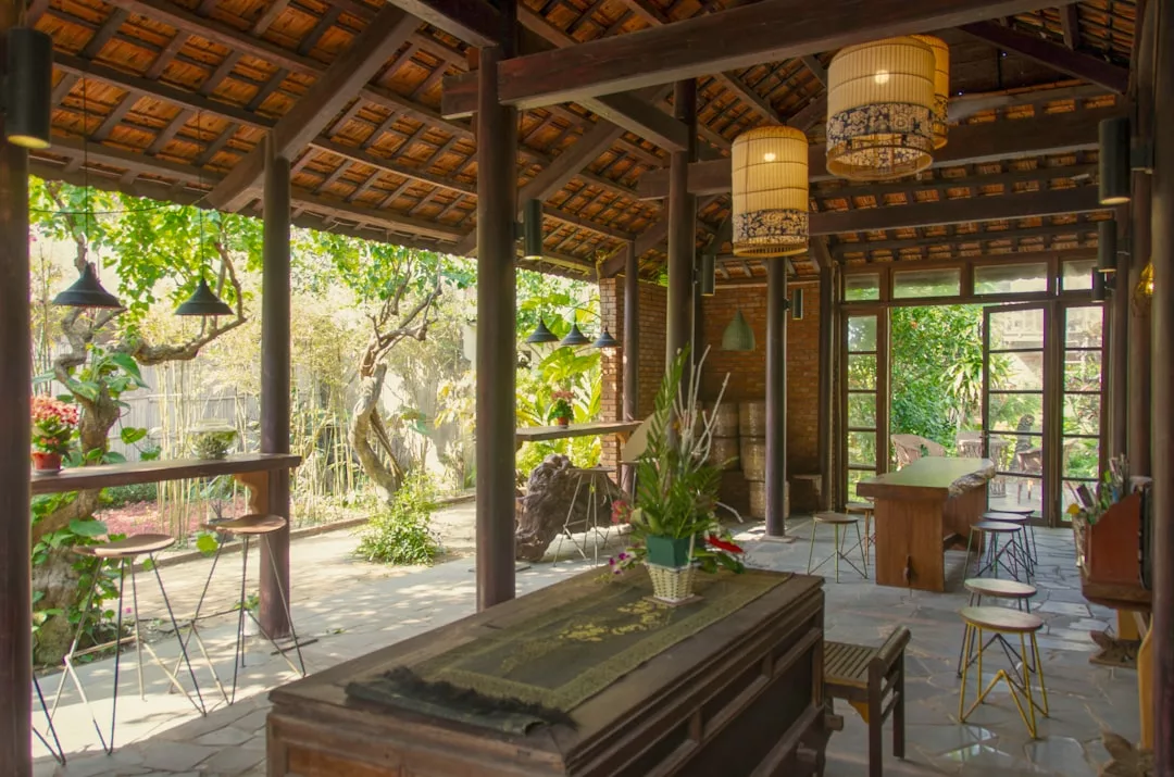 Comedor al aire libre con muebles de madera, lámparas colgantes y vigas a la vista, que ofrece un toque de lujo con vistas a un exuberante jardín a través de grandes ventanas y puertas abiertas, evocando la relajada elegancia de Mallorca.