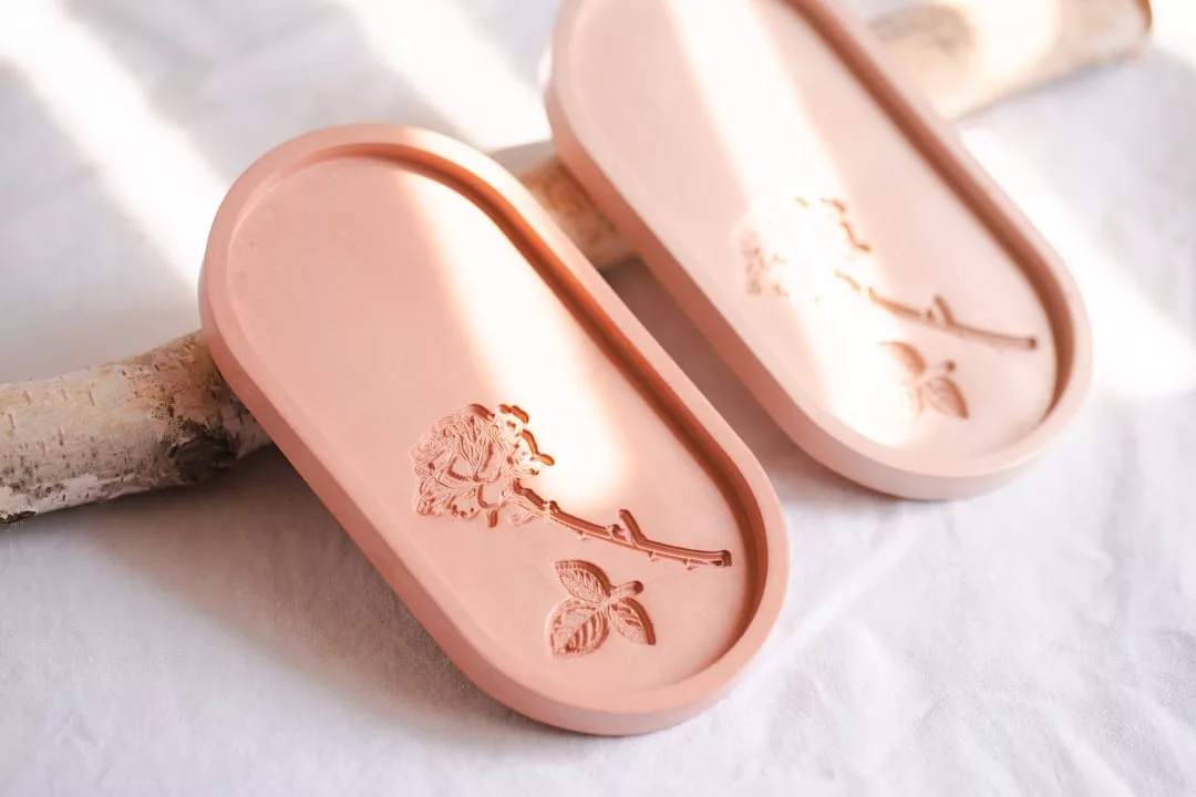Dos bandejas ovaladas de color rosa con un diseño de rosas en relieve descansan sobre una superficie blanca, parcialmente apoyadas en una rama de madera clara, evocando el relajado lujo del encanto costero de Mallorca.