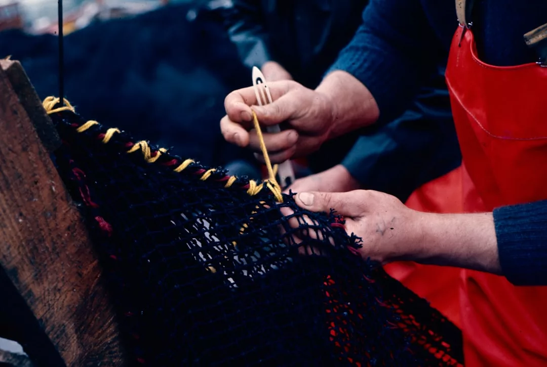 Dos personas en Mallorca reparan una red de pesca negra con hilo amarillo; una lleva un delantal rojo, y ambas usan sus manos para tejer la red, mostrando la artesanía tradicional en medio del sencillo lujo de la vida isleña.