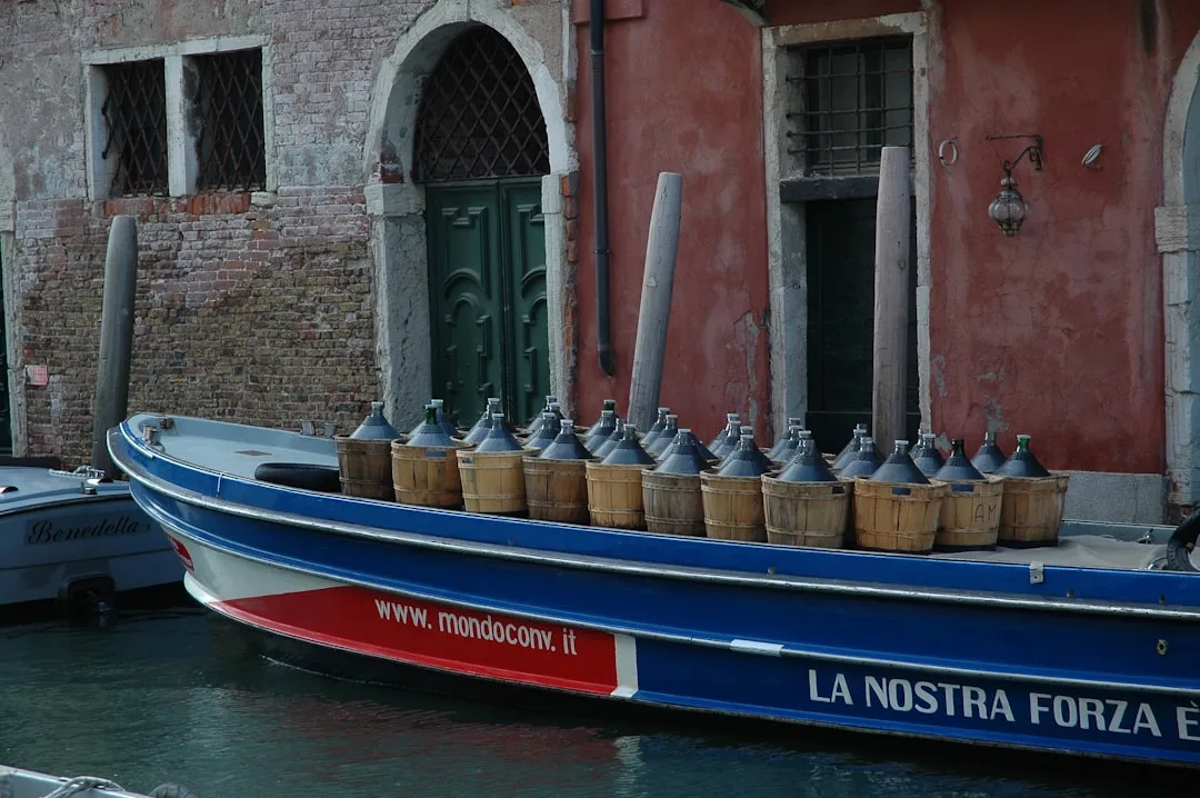 Un barco atracado junto a un canal de Venecia transporta varias garrafas de vino de gran tamaño en cestas de madera alineadas en la cubierta, evocando el lujo intemporal de Mallorca con un telón de fondo de edificios históricos.