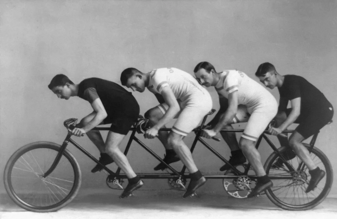 Cuatro hombres con uniforme de ciclistas montan en una bicicleta tándem vintage para cuatro personas en un estudio, evocando el espíritu de las rutas escénicas de Mallorca y la elegancia del lujo clásico.
