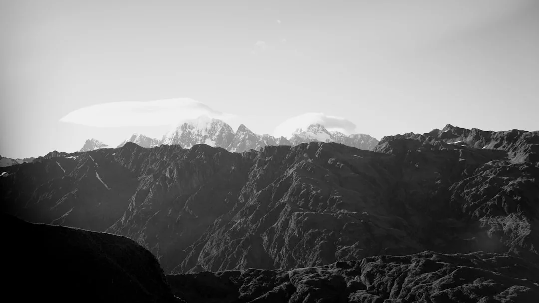 Fotografía en blanco y negro de escarpados picos montañosos en primer plano con lejanas montañas nevadas y arremolinadas formaciones nubosas, evocando los dramáticos paisajes de Mallorca con un toque de lujo.