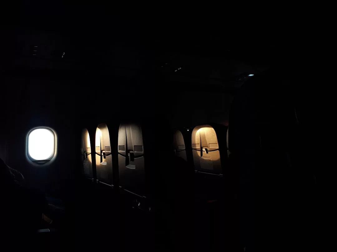 Una cabina de avión tenuemente iluminada, con varios asientos vacíos y una única ventanilla iluminada, evoca el tranquilo lujo de volar a Mallorca, donde la luz y la oscuridad se mezclan en un tranquilo contraste.