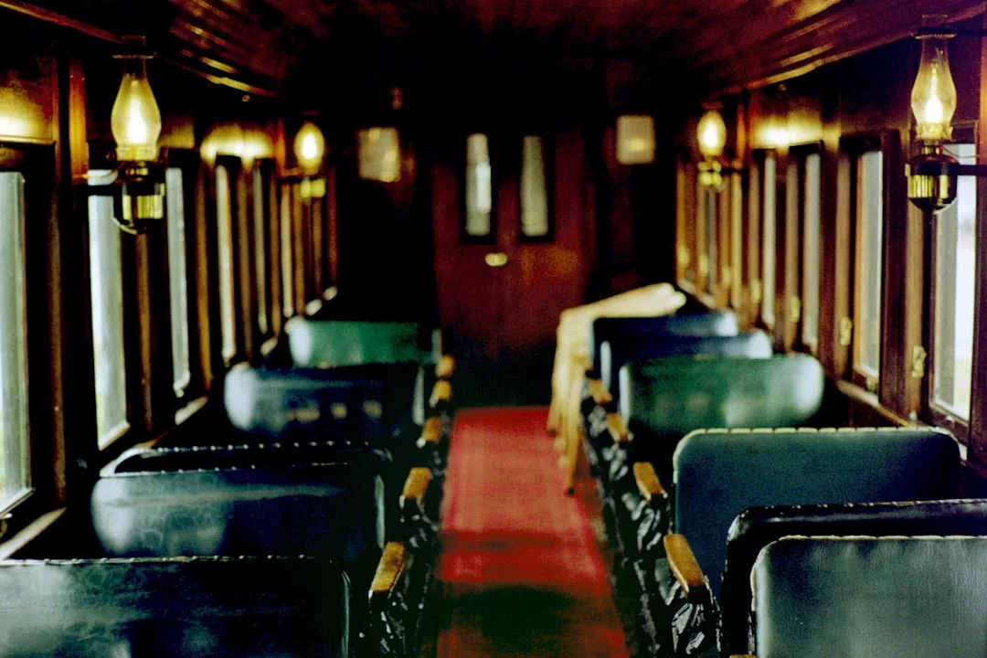 Interior de un vagón de tren de época en Mallorca, con asientos de cuero verde, paneles de madera, ventanas de cristal y lámparas de aceite en las paredes. Una alfombra roja recorre el pasillo central, añadiendo un toque de lujo a este nostálgico viaje.