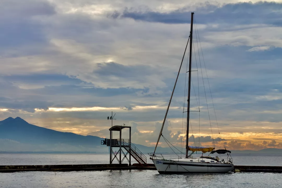 Un velero está atracado cerca de un muelle de madera con una pequeña caseta de observación, frente a un cielo nublado y montañas lejanas al atardecer, capturando el sereno lujo de la costa de Mallorca.