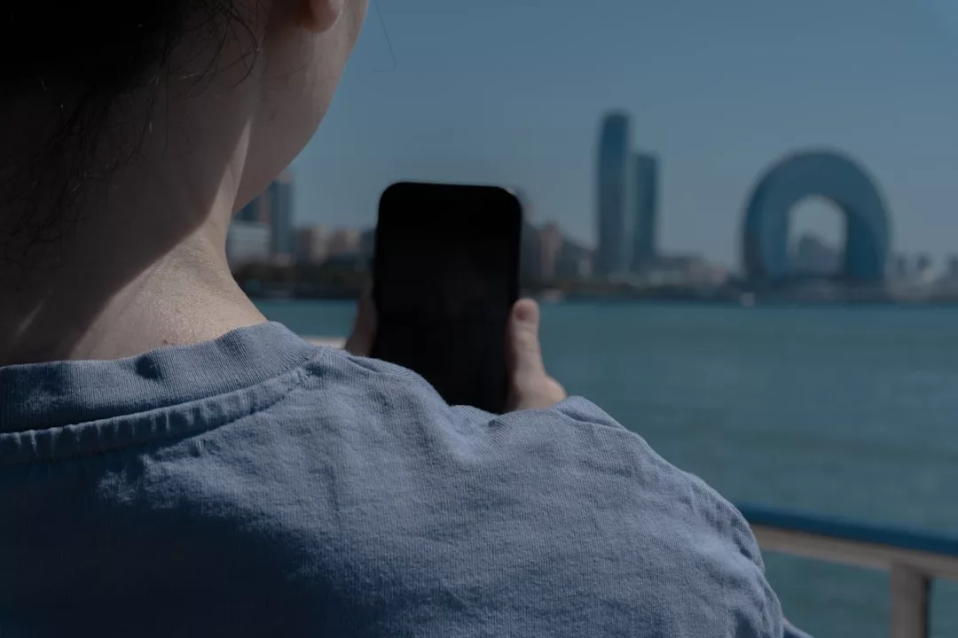 Una persona con camisa azul claro sostiene un smartphone frente a un paisaje urbano frente al mar con edificios modernos, captando el lujo y la elegancia que recuerdan a las impresionantes vistas costeras de Mallorca.