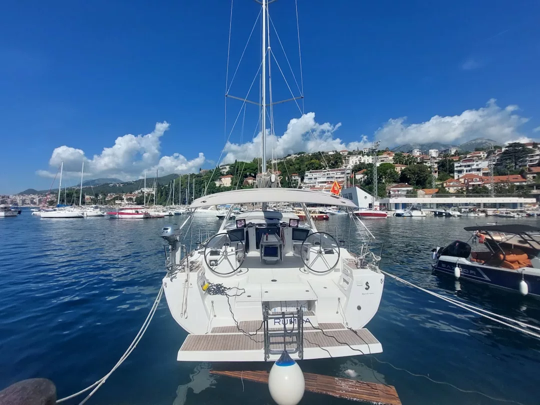 Un velero blanco está atracado en un puerto deportivo de Mallorca con varias otras embarcaciones, aguas tranquilas y edificios en las laderas bajo un cielo azul despejado, ofreciendo una escena de tranquilidad y lujo.