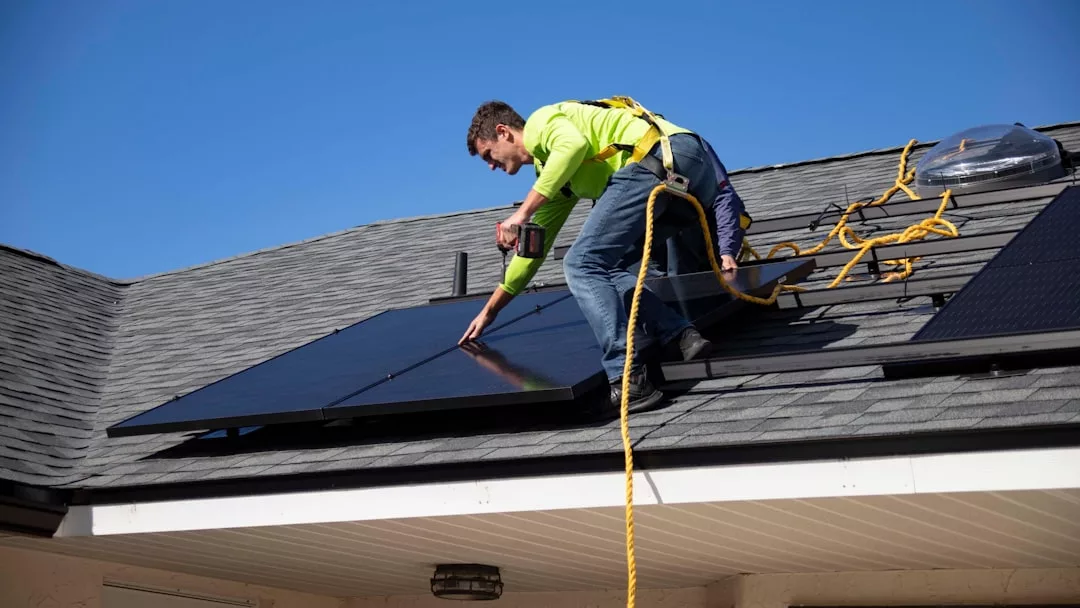 Un trabajador con equipo de seguridad instala paneles solares en un tejado residencial inclinado bajo un cielo azul despejado, aportando lujo sostenible a una vivienda de Mallorca.