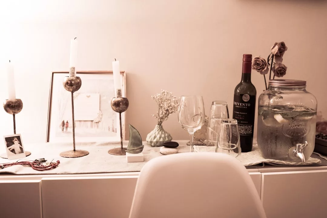 Una mesa con dos velas encendidas, un cuadro enmarcado, flores secas, copas de vino, una botella de vino tinto y un dispensador de vasos llenos de una bebida de limón aporta un aire de lujo a cualquier ambiente, perfecto para evocar las veladas en Mallorca.