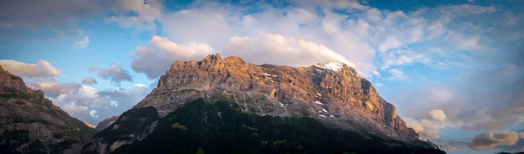 Una gran montaña con acantilados rocosos y manchas de nieve es iluminada por la luz del sol bajo un cielo con nubes dispersas, ofreciendo un toque de lujo que recuerda a los impresionantes paisajes de Mallorca.