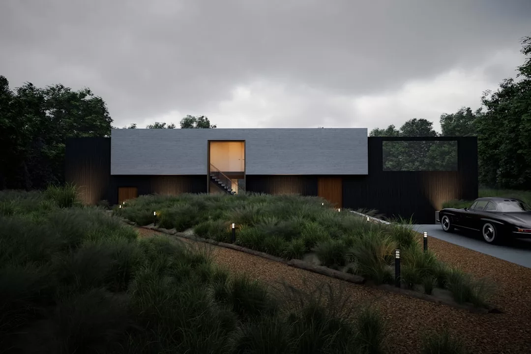Una casa moderna, rectangular, con paredes exteriores oscuras y grandes ventanales, rezuma lujo en medio de hierba alta y arbustos en Mallorca, con un camino de grava y un coche aparcado delante bajo un cielo nublado.